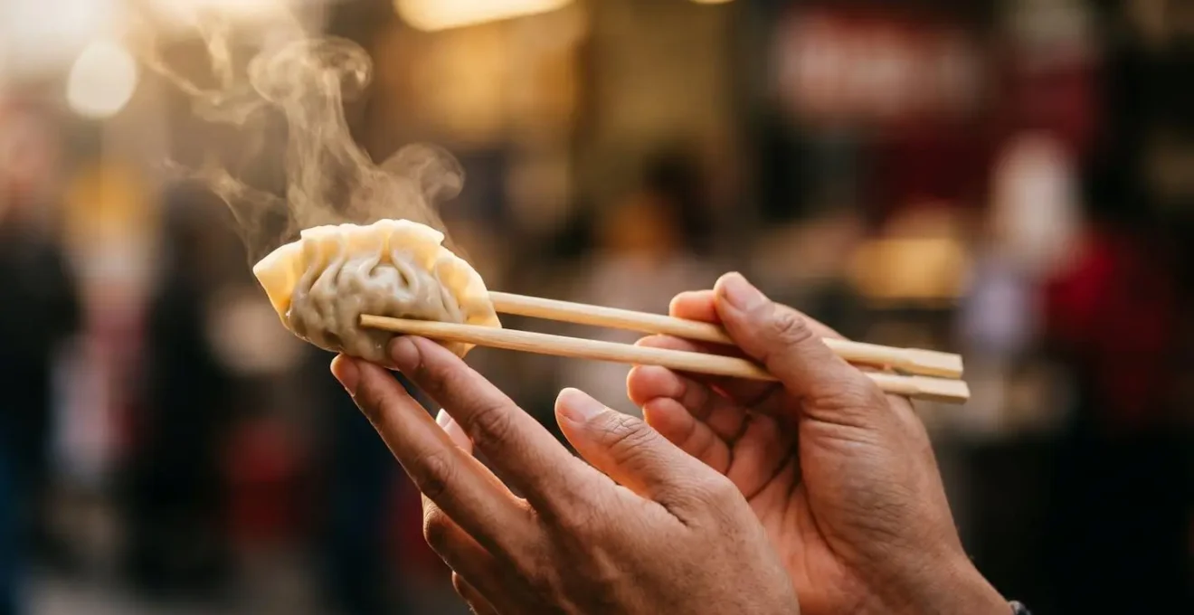 Close-up of hands holding a steaming dumpling with chopsticks against blurred market background showcasing food evaluation